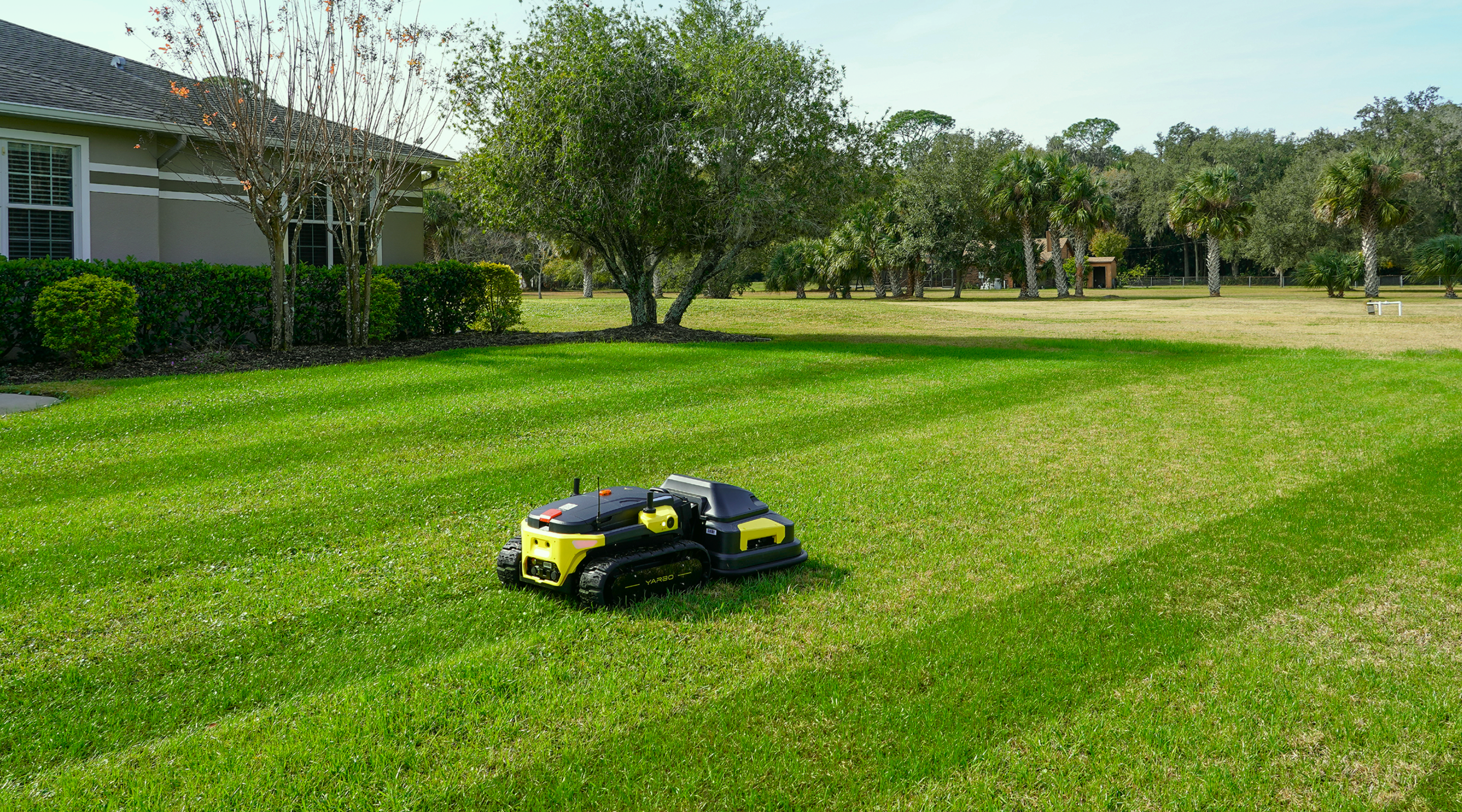 Yarbo robot lawn mower autonomously mowing a large open residential lawn with visible stripe pattern, surrounded by trees and landscaping