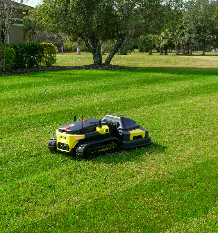 Yarbo robot lawn mower autonomously mowing a large open residential lawn with visible stripe pattern, surrounded by trees and landscaping